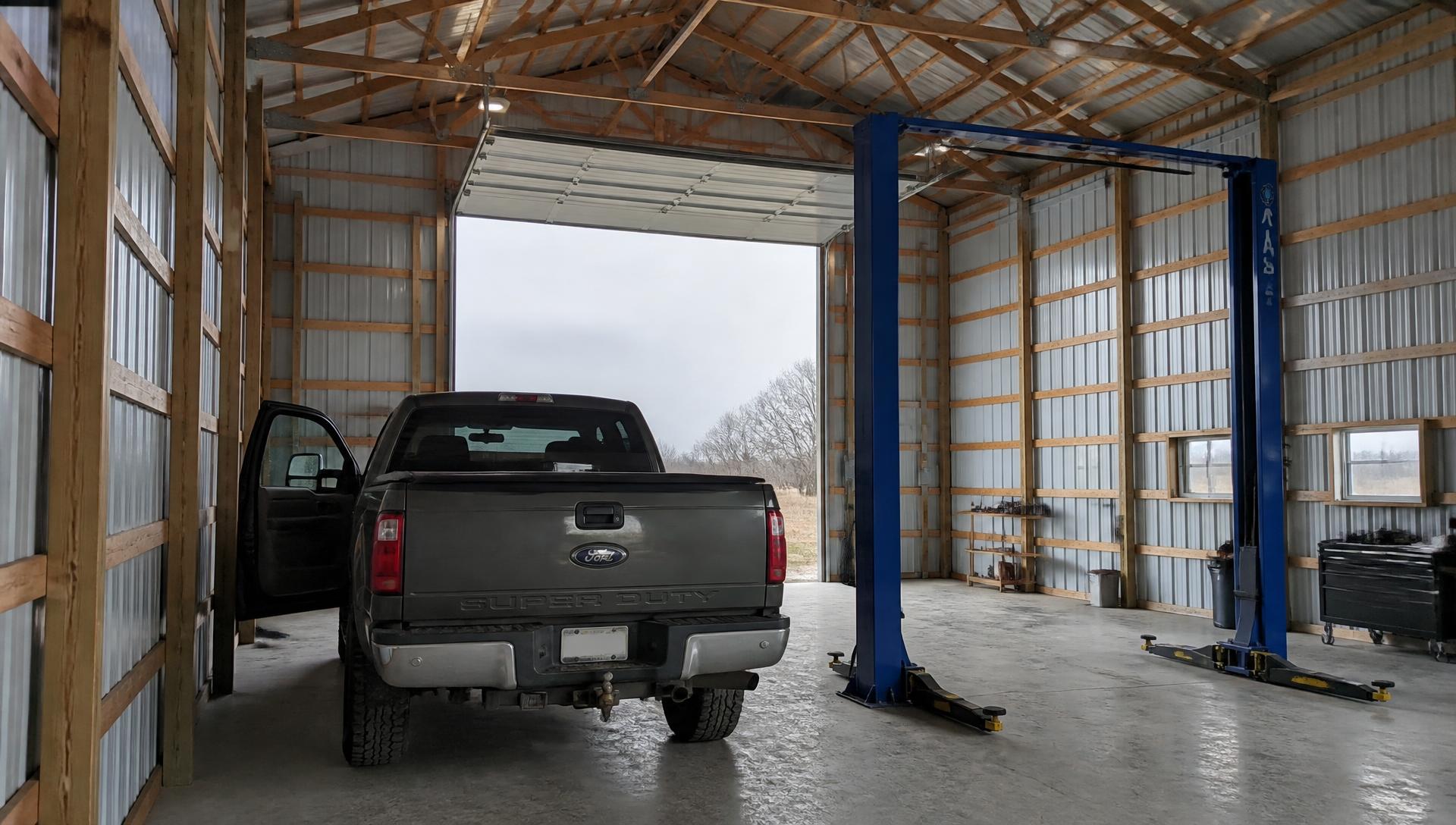 A full size pickup parked with its driver door open inside a tall post frame shop, leaving barely a walking gap beside an empty two post car lift